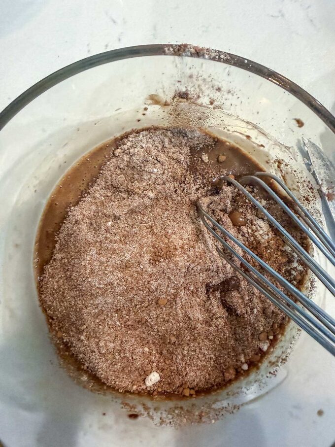 A glass bowl with cocoa powder and liquid being mixed with a metal whisk on a white surface, ready for baking preparation—perfect for crafting a delicious Keto Chocolate Mug Cake or any other easy Keto dessert.