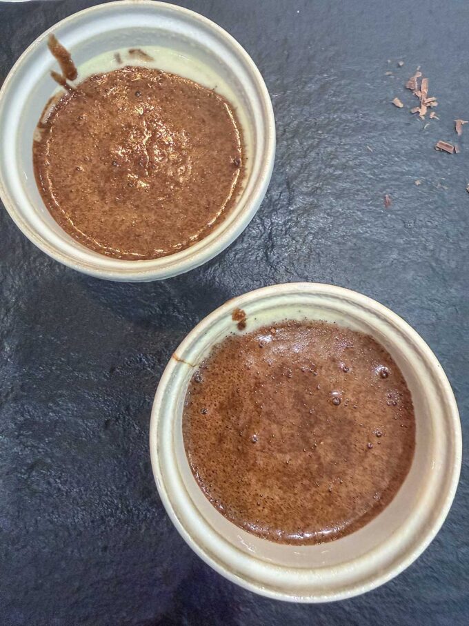 Two small ceramic bowls filled with rich, dark chocolate mousse sit on a black textured surface—a perfect keto dessert for chocolate lovers. Some chocolate shavings are scattered nearby, adding an elegant finishing touch.