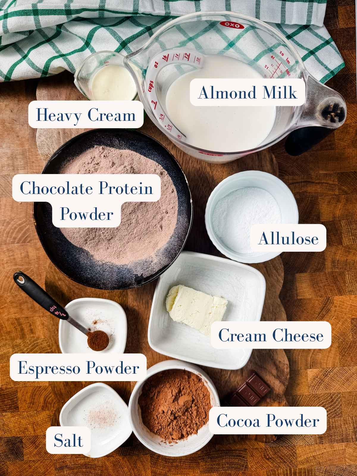 Top-down view of labeled ingredients in bowls and cups on a wooden surface, ready for making chocolate protein pudding: heavy cream, almond milk, chocolate protein powder, allulose, cream cheese, cocoa powder, espresso powder, and salt. A checkered cloth is in the background.